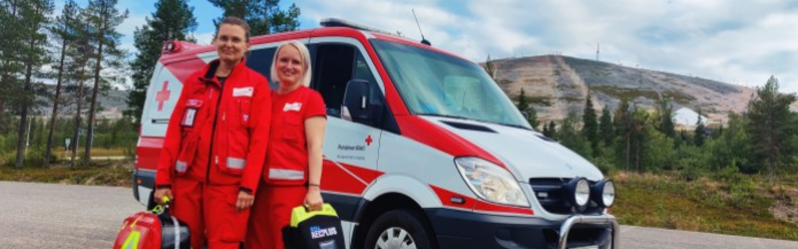 Kaksi Suomen Punaisen Ristin vapaaehtoista seisoo ambulanssin edessä tunturin juurella.