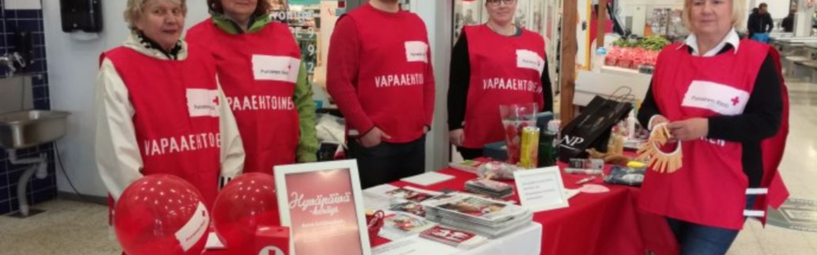 Joukko vapaaehtoisia pitää Suomen Punaisen Ristin pistettä kauppakeskuksessa. Vapaaehtoiset seisovat rivissä ja hymyilevät kameralle.