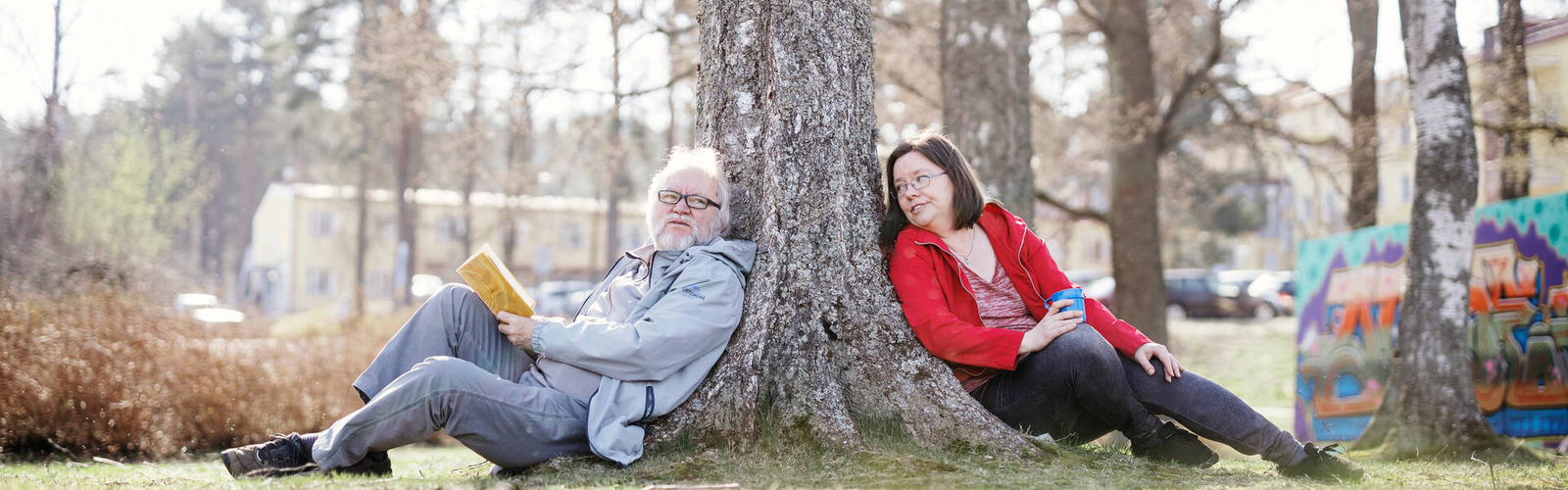 Kaksi henkilöä ulkona nojaa puunrunkoon ja hymyilee. 