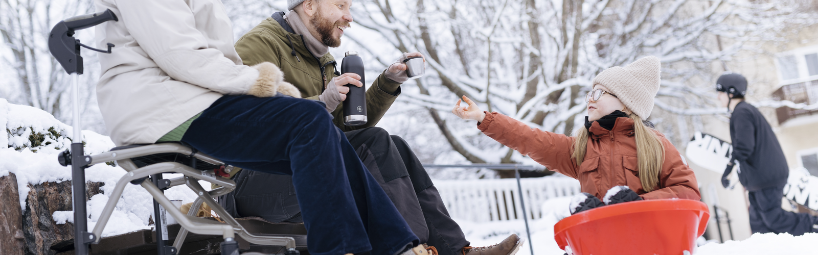 Lapsi istuu pulkassa ja ottaa isältään termosmukissa lämmintä juotavaa ja isän vieressä istuu vanhempi naishenkilö, kenties lapsen mummo