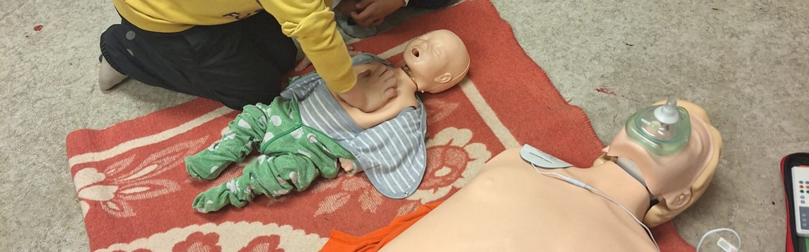 kuvassa kaksi lasta elvyttämässä baby-nukkea, lisäksi kuvassa anne-nukke ja defibrillaattori
