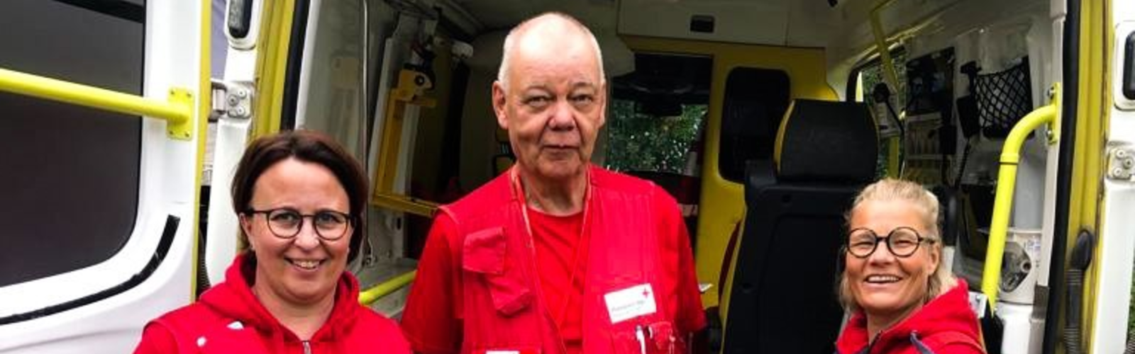 Kuvassa seisoo kolme ensiapuryhmäläistä ensiapu ambulanssin takana